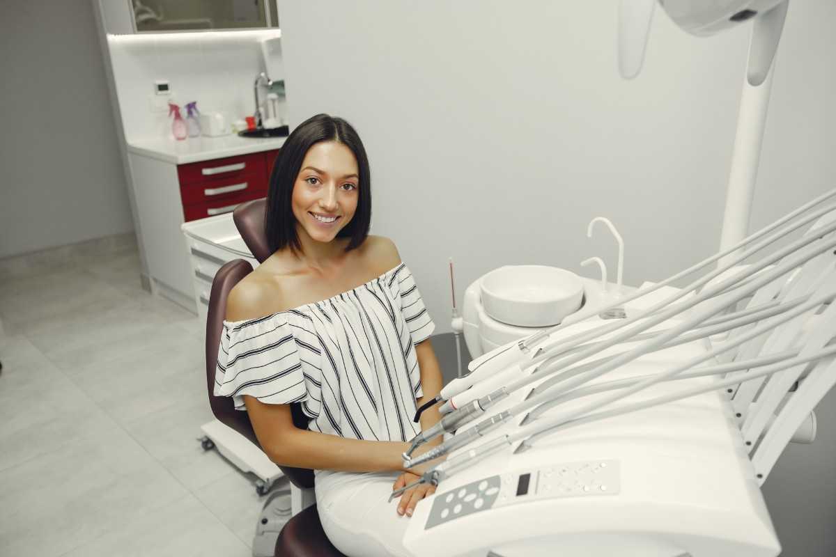 Smiling female patient sitting in a dental chair in a modern dental office, with dental instruments and equipment visible nearby.